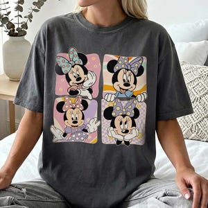 May include: Dark gray t-shirt featuring a four-panel design of Minnie Mouse. Each panel showcases a different pose of Minnie Mouse with a polka dot bow, set against a pastel background. The shirt is a comfortable, casual style.
