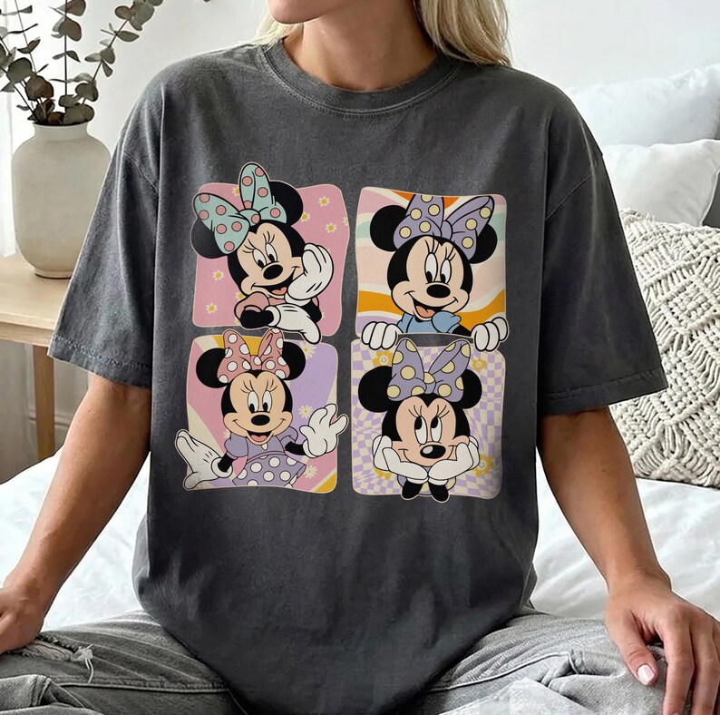 May include: Dark gray t-shirt featuring a four-panel design of Minnie Mouse. Each panel showcases a different pose of Minnie Mouse with a polka dot bow, set against a pastel background. The shirt is a comfortable, casual style.