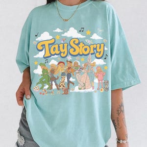 K&ouml;nnte beinhalten: Ein hellblaues T-Shirt mit einem Grafikdruck, der den Text Tay Story in gelben Buchstaben zeigt. Das Design umfasst eine Gruppe illustrierter Figuren in verschiedenen Outfits, umgeben von wei&szlig;en Wolken, gelben Sternen und Noten.