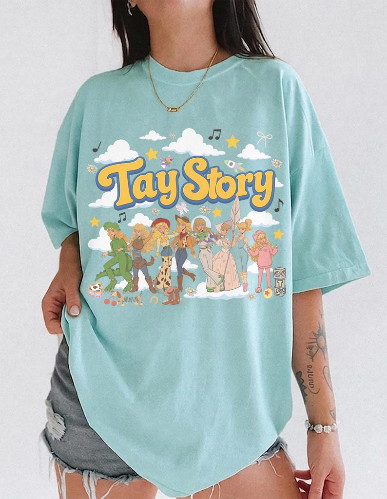K&ouml;nnte beinhalten: Ein hellblaues T-Shirt mit einem Grafikdruck, der den Text Tay Story in gelben Buchstaben zeigt. Das Design umfasst eine Gruppe illustrierter Figuren in verschiedenen Outfits, umgeben von wei&szlig;en Wolken, gelben Sternen und Noten.