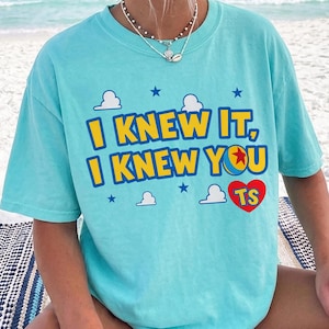 May include: A light turquoise short-sleeve t-shirt featuring the yellow and blue text I KNEW IT, I KNEW YOU with a red heart containing the letters TS. The design includes small white clouds and dark blue stars printed on the front of the shirt.