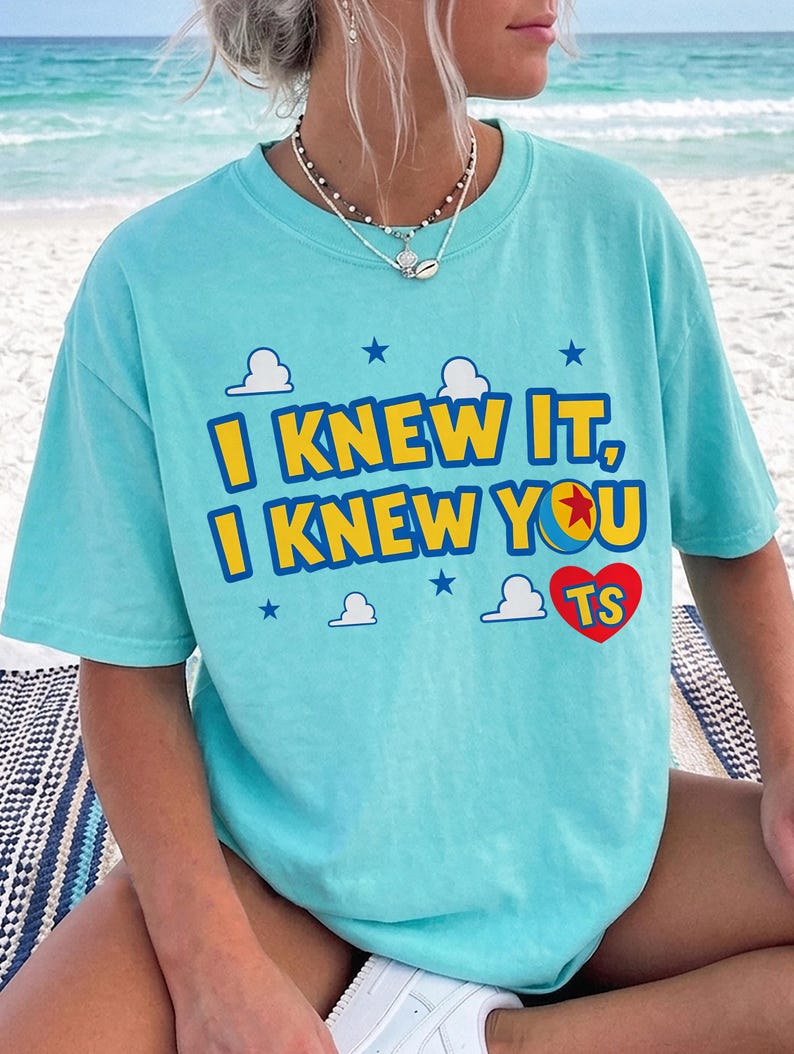 May include: A light turquoise short-sleeve t-shirt featuring the yellow and blue text I KNEW IT, I KNEW YOU with a red heart containing the letters TS. The design includes small white clouds and dark blue stars printed on the front of the shirt.