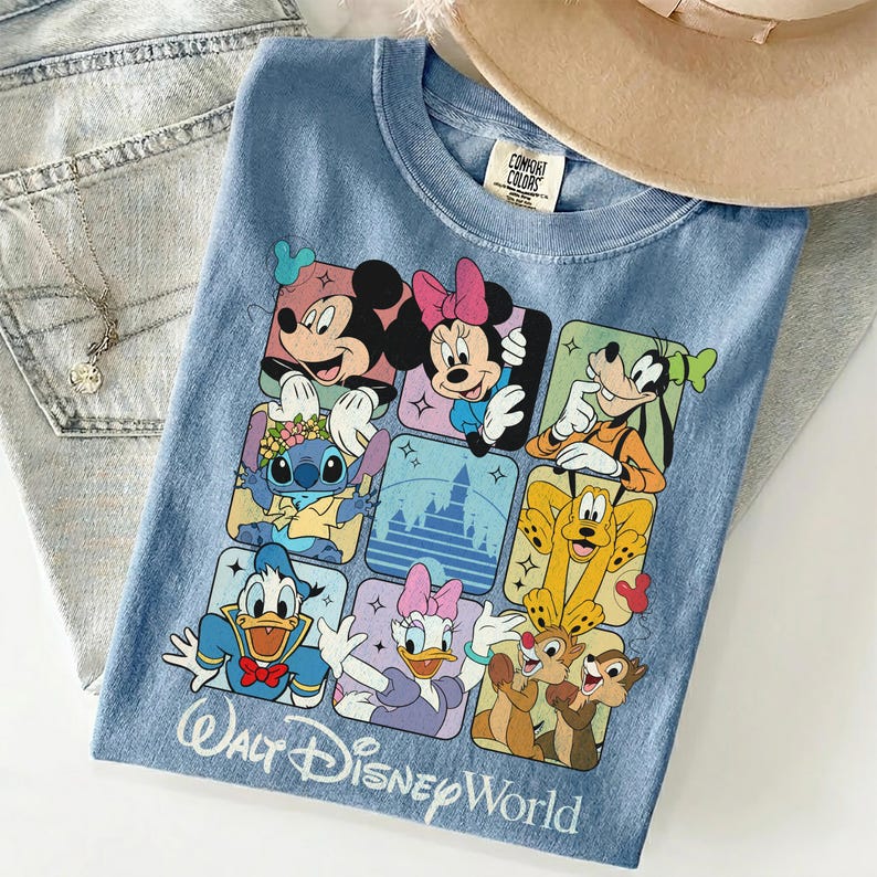 May include: Light blue t-shirt featuring a colorful graphic of Disney characters, including Mickey Mouse, Minnie Mouse, and Goofy. The shirt has the text "Walt Disney World" in a script font. A beige hat and a silver necklace are also visible.