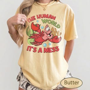 May include: Butter-colored t-shirt featuring a cartoon crab with the text "THE HUMAN WORLD IT'S A MESS". The crab is red with a smiling expression, surrounded by bubbles and seaweed. The shirt is a casual, comfortable style.