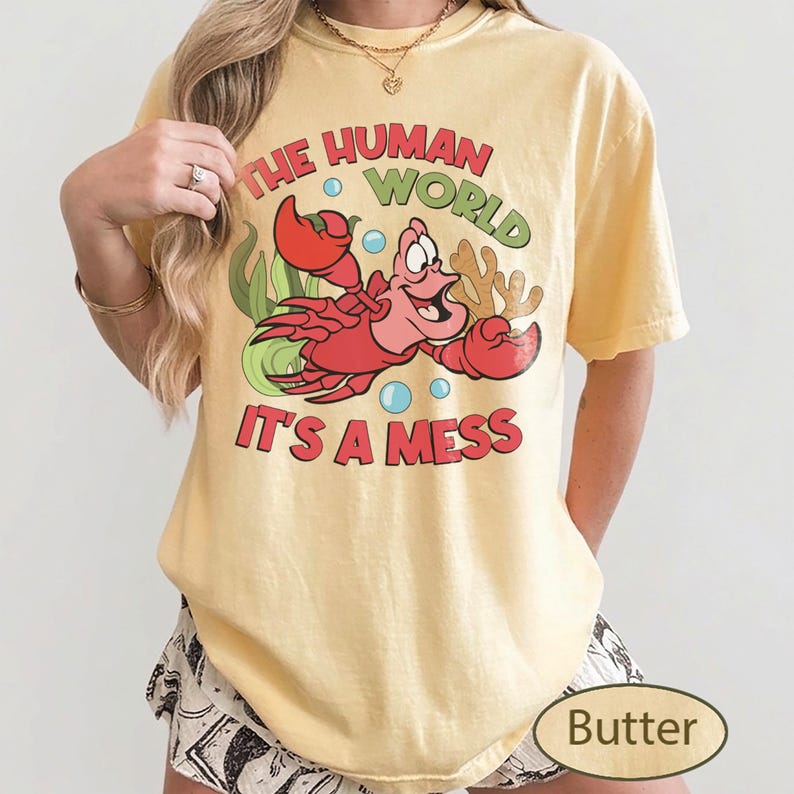 May include: Butter-colored t-shirt featuring a cartoon crab with the text "THE HUMAN WORLD IT'S A MESS". The crab is red with a smiling expression, surrounded by bubbles and seaweed. The shirt is a casual, comfortable style.