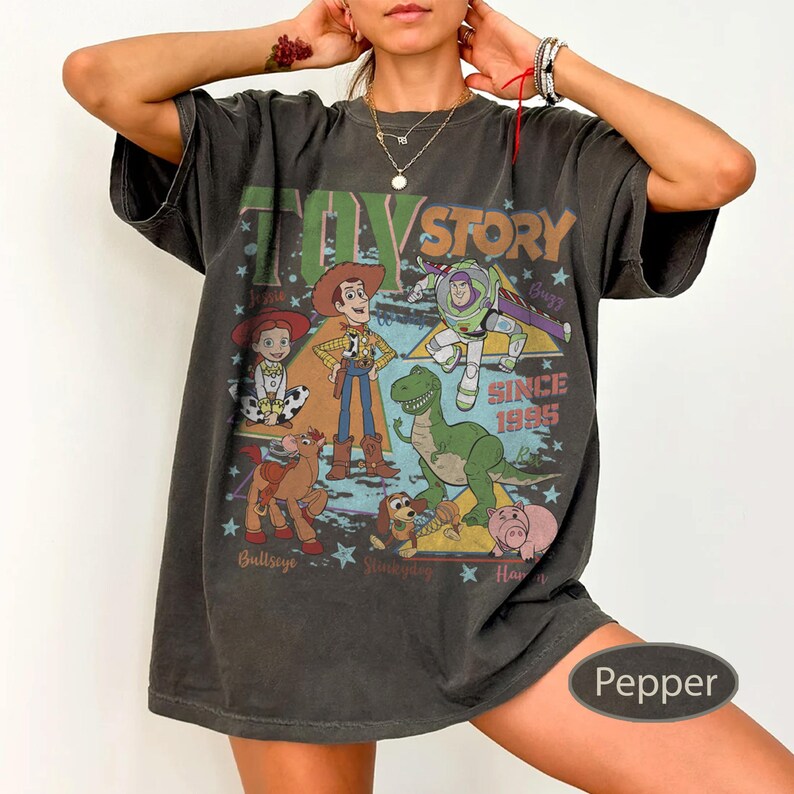 Puede incluir: Camiseta color pimienta con un dise&ntilde;o gr&aacute;fico de personajes de la pel&iacute;cula Toy Story. El dise&ntilde;o incluye a Woody, Buzz Lightyear y otros personajes, con el texto "TOY STORY SINCE 1995". La camiseta tiene un aspecto vintage.