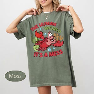 May include: Moss green t-shirt featuring a cartoon image of a red crab with the text "THE HUMAN WORLD IT'S A MESS". The graphic includes bubbles, seaweed, and coral. The shirt is a relaxed fit.