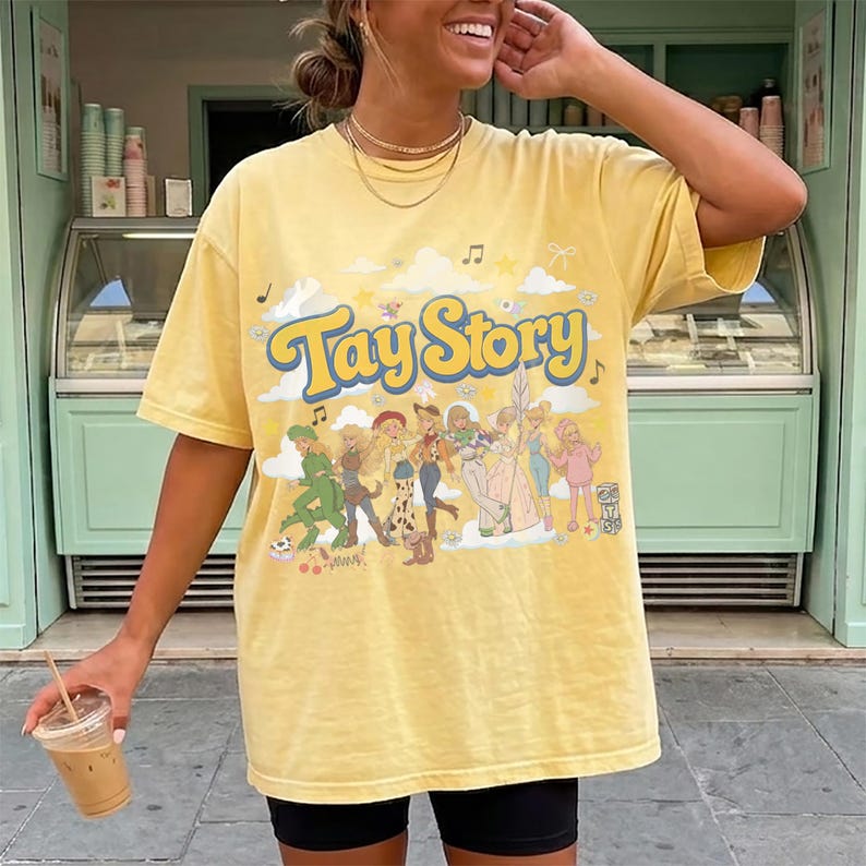 K&ouml;nnte beinhalten: Ein blassgelbes Kurzarm-T-Shirt mit dem Schriftzug Tay Story in blauen Buchstaben. Die Grafik zeigt eine Gruppe illustrierter Figuren in verschiedenen Outfits, dazu Noten, Sterne und Wolken, die mittig auf der Brust aufgedruckt sind.