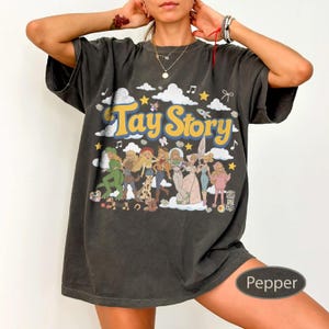 K&ouml;nnte beinhalten: Ein anthrazitfarbenes Kurzarm-T-Shirt mit dem gelben Schriftzug Tay Story. Die Grafik zeigt eine Gruppe illustrierter Figuren in verschiedenen Outfits, umgeben von wei&szlig;en Wolken, gelben Sternen und Noten auf dunklem Hintergrund.