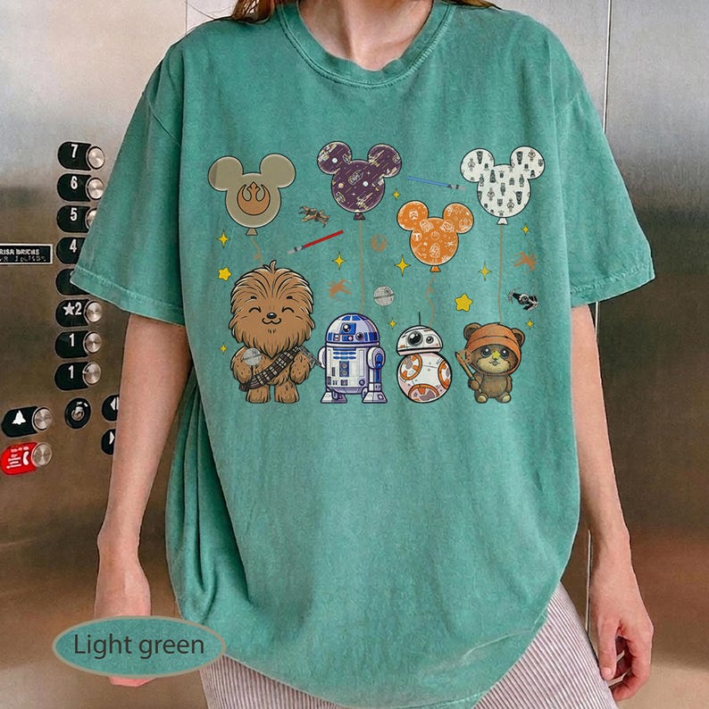 May include: Light green t-shirt featuring cartoon characters from Star Wars and Mickey Mouse. The design includes Chewbacca, R2-D2, BB-8, an Ewok, and Mickey Mouse balloons. The shirt has a relaxed fit.