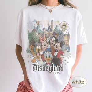 May include: A white short-sleeve t-shirt featuring a colorful graphic print of Mickey Mouse, Minnie Mouse, Donald Duck, Goofy, and Pluto in front of a Disneyland castle. The text Disneyland Est. 1955 is printed in black below the character illustration.