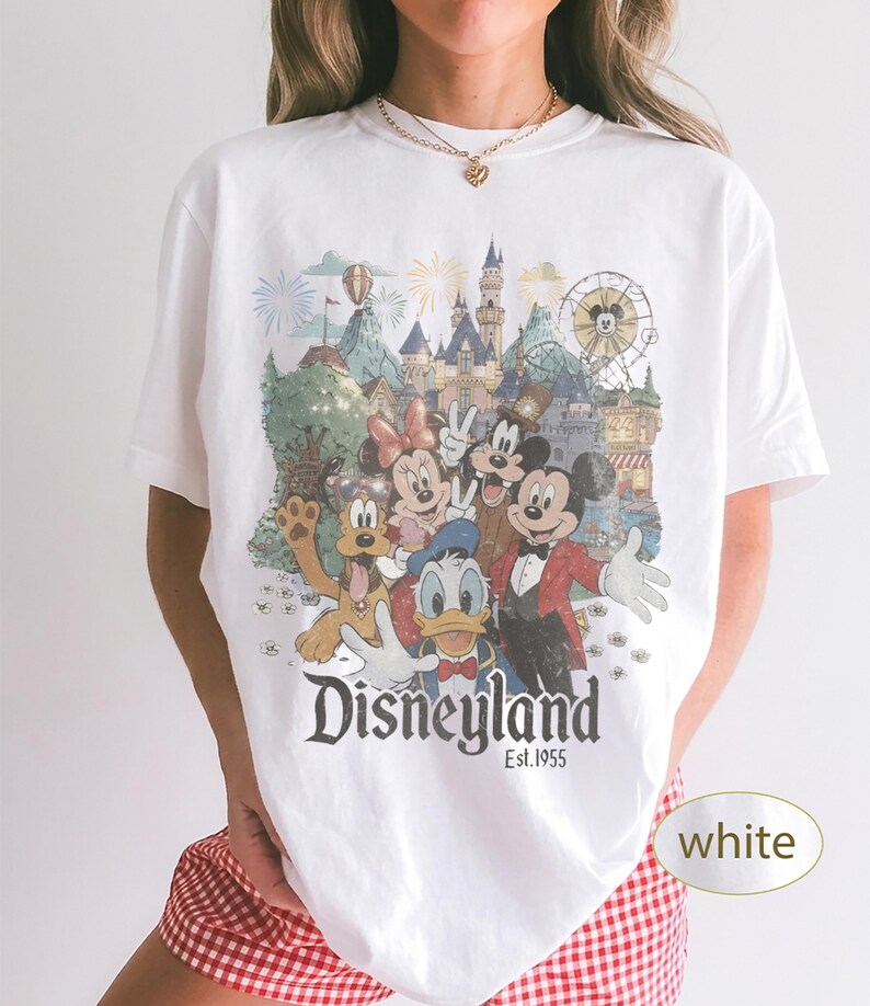 May include: A white short-sleeve t-shirt featuring a colorful graphic print of Mickey Mouse, Minnie Mouse, Donald Duck, Goofy, and Pluto in front of a Disneyland castle. The text Disneyland Est. 1955 is printed in black below the character illustration.