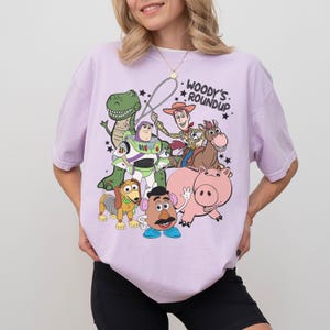 May include: A light purple t-shirt featuring a graphic of characters from the animated film Toy Story. The design includes Woody, Buzz Lightyear, Rex, Hamm, Mr. Potato Head, Slinky Dog, and a horse. The text "Woody's Roundup" is also included.