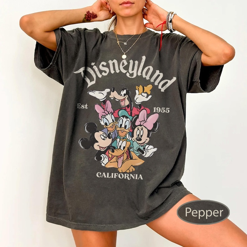 May include: A dark gray t-shirt featuring the word "Disneyland" above a graphic of Disney characters, including Mickey and Minnie Mouse, Donald Duck, and Goofy. The shirt also includes the text "Est 1955" and "California".