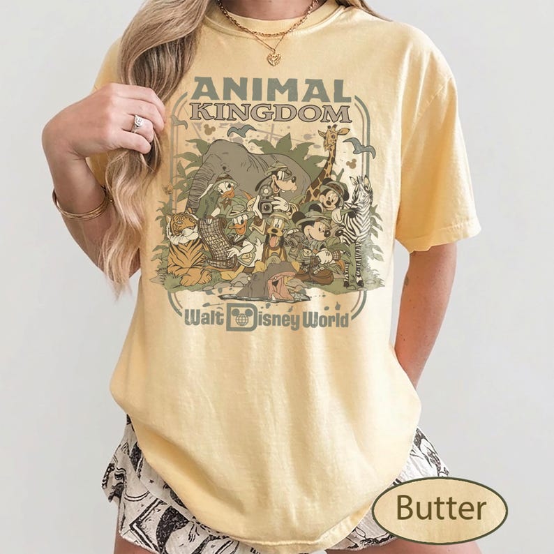 May include: Butter-colored t-shirt featuring a vintage-style graphic of Disney's Animal Kingdom. The design includes various animal characters and the text "ANIMAL KINGDOM" and "Walt Disney World" in a retro font. The shirt has short sleeves.
