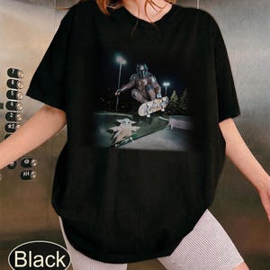 May include: A black short-sleeved t-shirt featuring a graphic print of a character in metallic armor performing a trick on a skateboard above a small green-robed creature on a separate skateboard in a concrete skate park at night.