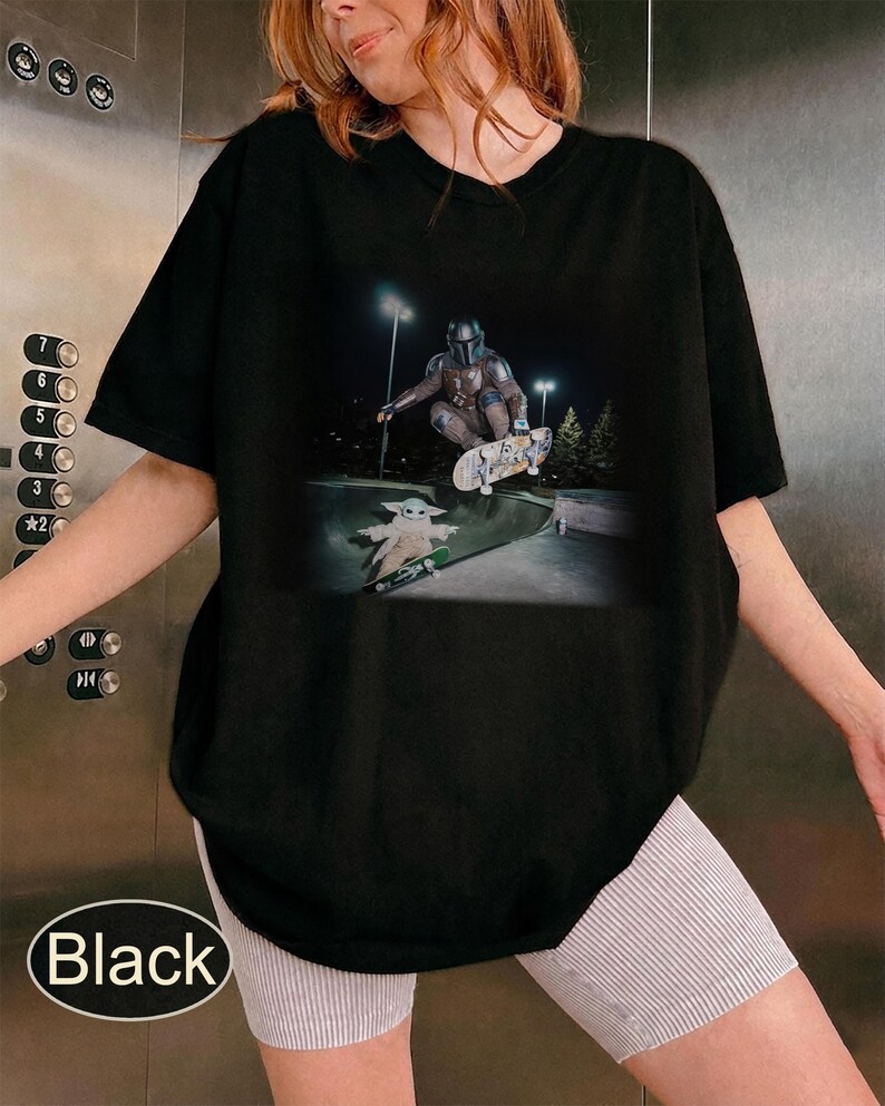 May include: A black short-sleeved t-shirt featuring a graphic print of a character in metallic armor performing a trick on a skateboard above a small green-robed creature on a separate skateboard in a concrete skate park at night.