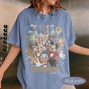 May include: A blue short-sleeve t-shirt featuring a graphic print of Mickey Mouse, Minnie Mouse, Donald Duck, Goofy, and Pluto in front of a castle. The text Disneyland Est. 1955 is printed in gold below the characters, with fireworks in the background.