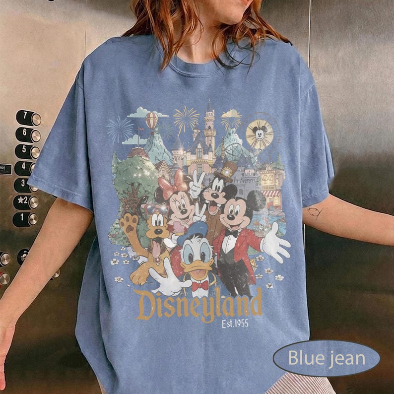 May include: A blue short-sleeve t-shirt featuring a graphic print of Mickey Mouse, Minnie Mouse, Donald Duck, Goofy, and Pluto in front of a castle. The text Disneyland Est. 1955 is printed in gold below the characters, with fireworks in the background.