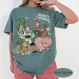 May include: Blue Spruce t-shirt featuring a graphic of Toy Story characters, including Woody, Buzz Lightyear, and Rex. The text "Woody's Roundup" is printed above the characters. The shirt is a relaxed fit.