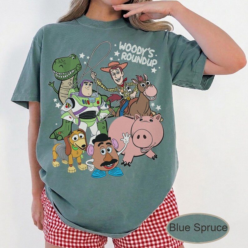 May include: Blue Spruce t-shirt featuring a graphic of Toy Story characters, including Woody, Buzz Lightyear, and Rex. The text "Woody's Roundup" is printed above the characters. The shirt is a relaxed fit.