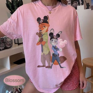 Pu&ograve; includere: T-shirt rosa chiaro con un'illustrazione a cartoni animati di Nick Wilde e Judy Hopps di Zootopia, con orecchie di Topolino. La maglietta ha maniche corte e scollo tondo. La parola "Blossom" &egrave; stampata in basso a sinistra.