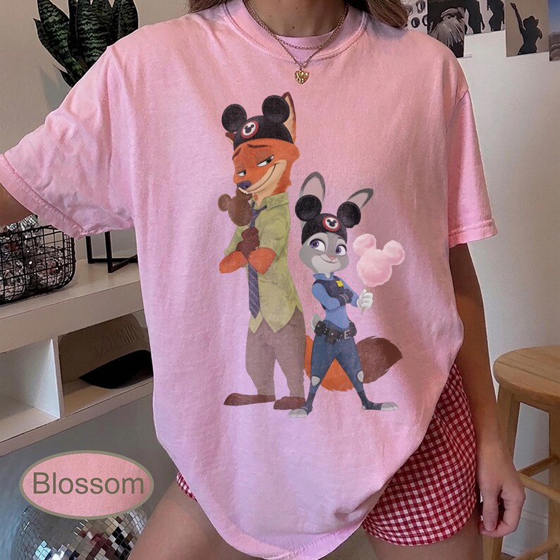 Pu&ograve; includere: T-shirt rosa chiaro con un'illustrazione a cartoni animati di Nick Wilde e Judy Hopps di Zootopia, con orecchie di Topolino. La maglietta ha maniche corte e scollo tondo. La parola "Blossom" &egrave; stampata in basso a sinistra.