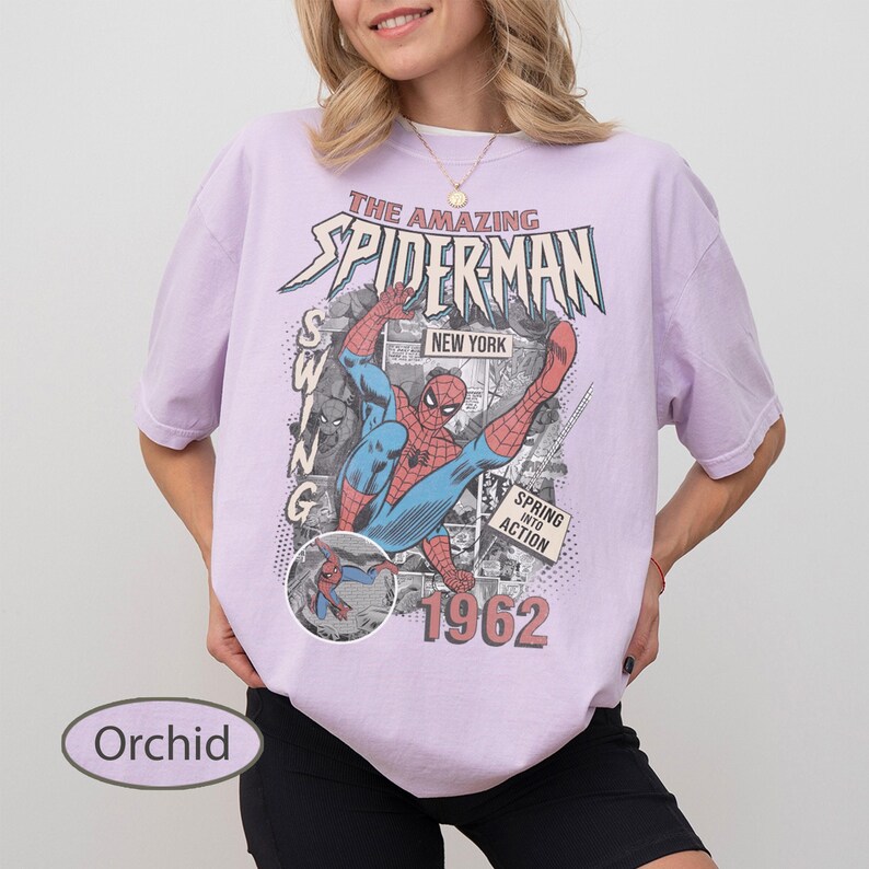 May include: Orchid-colored t-shirt featuring a vintage comic book graphic of Spider-Man in action. The design includes the text "The Amazing Spider-Man," "New York," and "1962." The shirt has short sleeves and a relaxed fit.