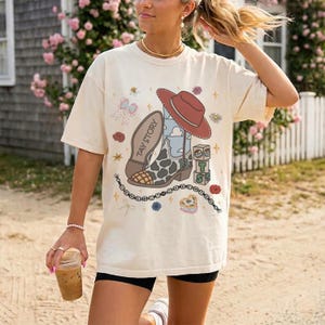 May include: An off-white short-sleeve t-shirt featuring a graphic print of a cowboy boot with a cow print toe, a red hat, alphabet blocks with the letters T, S, and a heart, a bow, flowers, stars, and a beaded necklace with the text TAY STORY.
