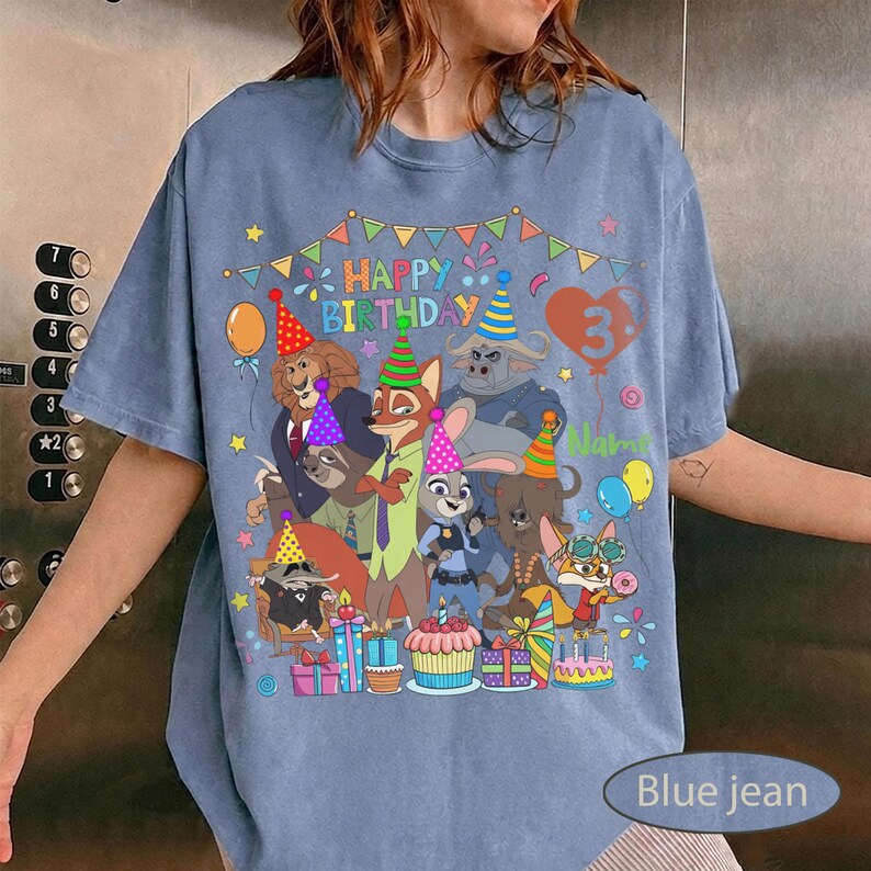 May include: Blue jean colored t-shirt with a colorful graphic featuring cartoon characters and the text "HAPPY BIRTHDAY" and the number "3". The design includes balloons, presents, and a cake.