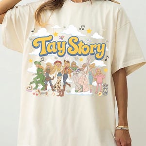 K&ouml;nnte beinhalten: Ein elfenbeinfarbenes T-Shirt mit einem Grafikdruck, der den Text "Tay Story" in gelben Buchstaben mit blauem Umriss zeigt. Das Design enth&auml;lt Illustrationen mehrerer blonder Figuren in verschiedenen Outfits, Wolken, Sterne und Musiknoten.