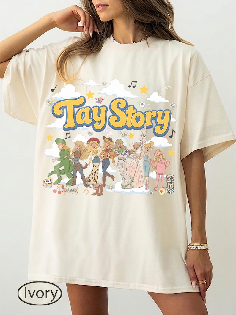 K&ouml;nnte beinhalten: Ein elfenbeinfarbenes T-Shirt mit einem Grafikdruck, der den Text "Tay Story" in gelben Buchstaben mit blauem Umriss zeigt. Das Design enth&auml;lt Illustrationen mehrerer blonder Figuren in verschiedenen Outfits, Wolken, Sterne und Musiknoten.