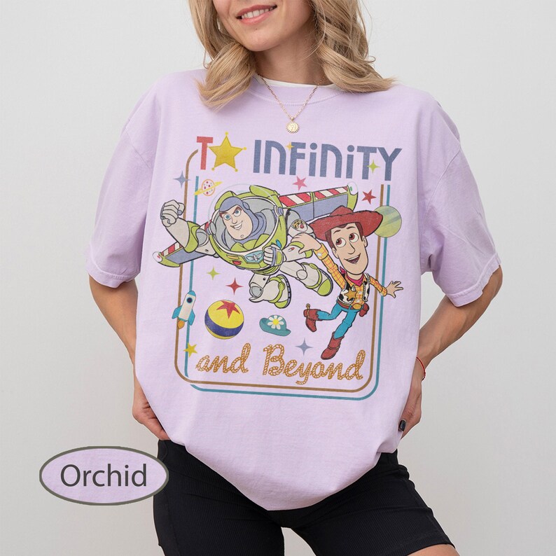 May include: Orchid-colored t-shirt featuring a graphic of Buzz Lightyear and Woody from Toy Story, with the text "To Infinity and Beyond." The design includes a rocket, stars, and a planet. The shirt is a relaxed fit.