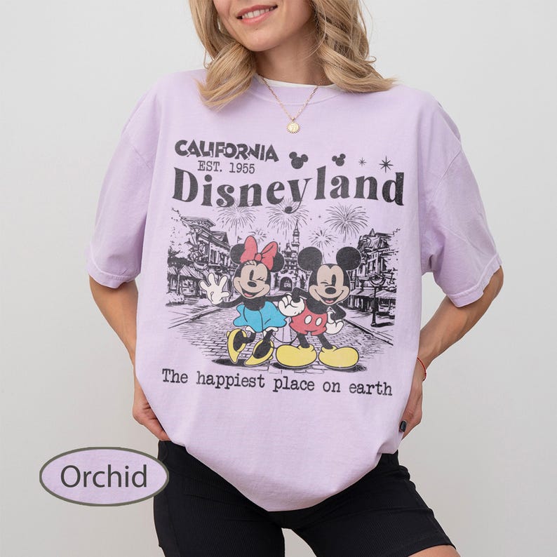 May include: Orchid-colored t-shirt featuring a Disneyland graphic. The design includes Mickey and Minnie Mouse, the text "California Est. 1955 Disneyland," and the phrase "The happiest place on earth."