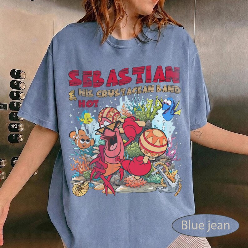 以下が含まれることがあります： 青いジーンズ色のTシャツで、セバスチャンというカニが他の海の生き物たちとマラカスを演奏しているグラフィックプリントが施されています。テキストには「SEBASTIAN & HIS CRUSTACEAN BAND」と書かれています。デザインにはサンゴ、錨、様々な魚のキャラクターが含まれています。