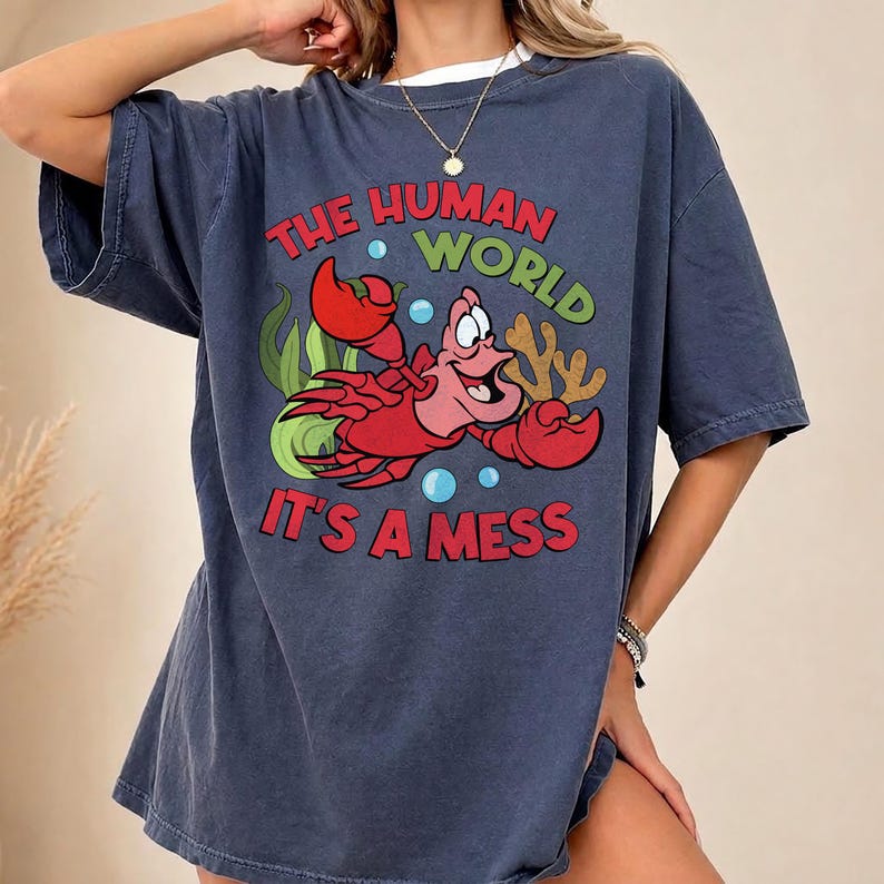 May include: A navy blue t-shirt featuring a cartoon image of a red crab with the text "THE HUMAN WORLD IT'S A MESS" in red and green. The shirt has a relaxed fit and a white undershirt is visible.