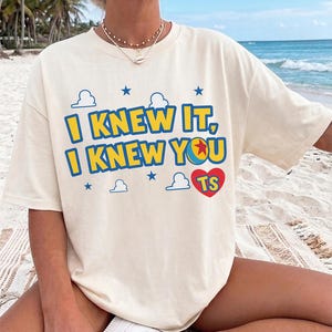 May include: A cream-colored short-sleeve t-shirt featuring the blue and yellow text I KNEW IT, I KNEW YOU with a colorful ball graphic, blue stars, white cloud outlines, and a red heart containing the letters TS printed on the front.