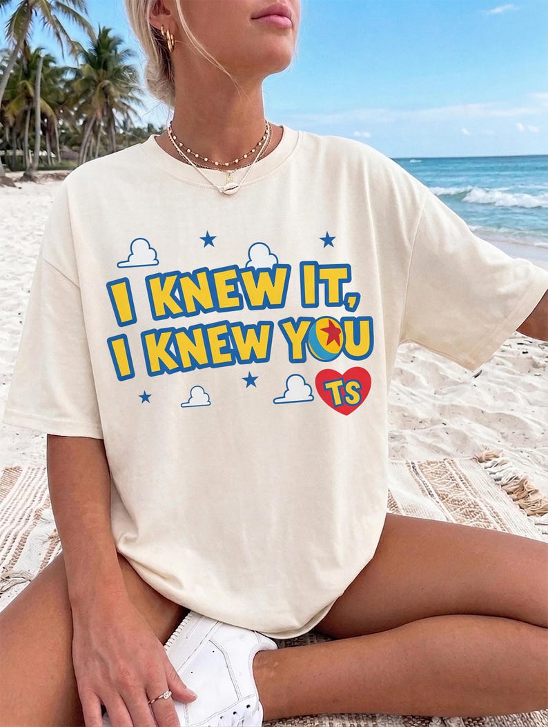May include: A cream-colored short-sleeve t-shirt featuring the blue and yellow text I KNEW IT, I KNEW YOU with a colorful ball graphic, blue stars, white cloud outlines, and a red heart containing the letters TS printed on the front.