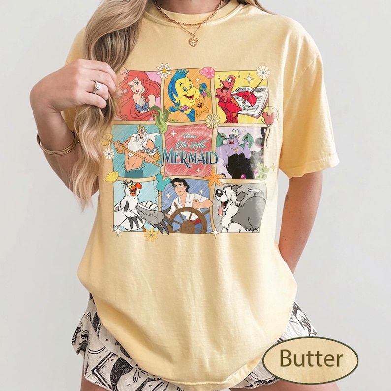 May include: Pale yellow t-shirt with a graphic featuring characters from The Little Mermaid. The design includes Ariel, Flounder, Sebastian, and other characters in a grid. The shirt has short sleeves and a loose fit.