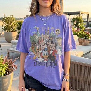 May include: A purple short-sleeve t-shirt featuring a graphic print of Disney characters including Mickey Mouse, Minnie Mouse, Donald Duck, and Pluto in front of a castle. The text Disneyland Est. 1955 is printed in dark gray below the characters.