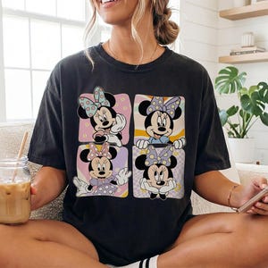 May include: Dark gray t-shirt featuring a four-panel design of Minnie Mouse in various poses. Each panel has a different background color and pattern, with Minnie wearing different outfits and bows. The shirt is a casual, oversized fit.