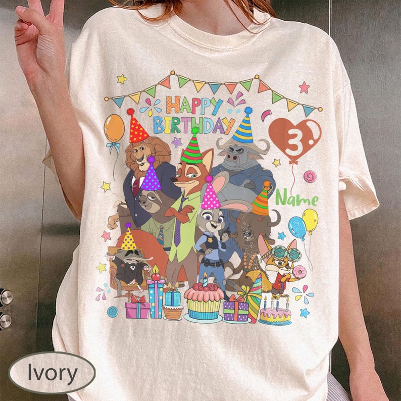 May include: Ivory-colored t-shirt featuring a colorful birthday design with cartoon characters, balloons, and the text "Happy Birthday." The number "3" is displayed, along with space for a name. Party hats and gifts are also included.
