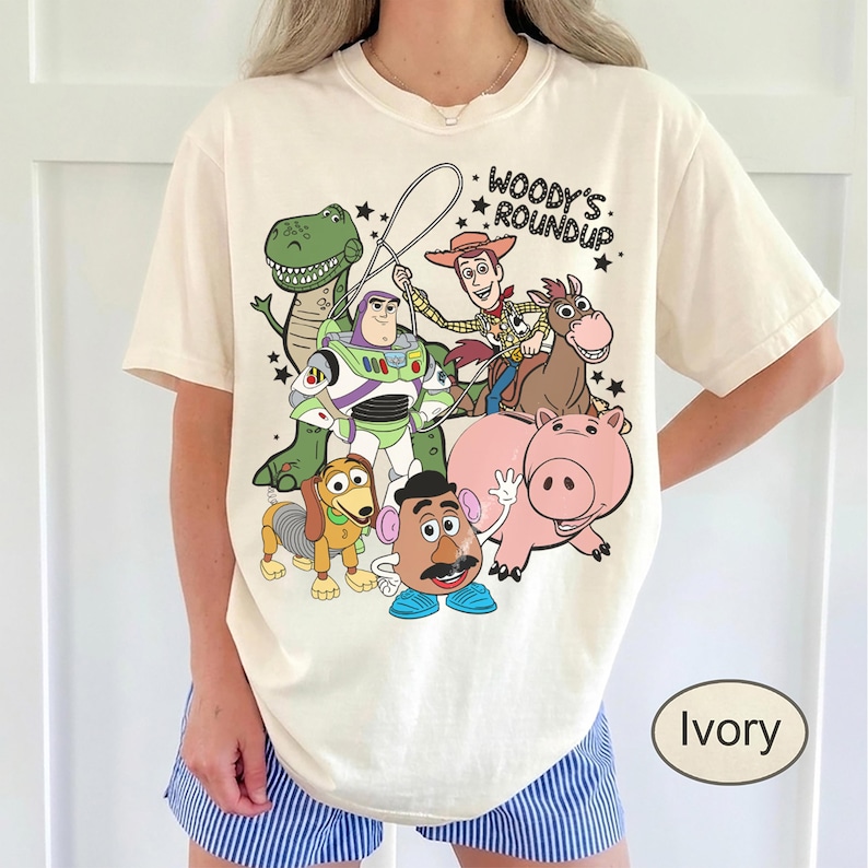 May include: Ivory-colored t-shirt featuring a graphic of characters from the animated film "Toy Story." The design includes Woody, Buzz Lightyear, Rex, Hamm, Slinky Dog, Mr. Potato Head, and a horse. Text above the characters reads "Woody's Roundup."