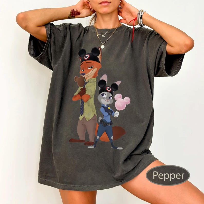 Pu&ograve; includere: T-shirt grigio scuro con un'illustrazione a cartoni animati di Nick Wilde e Judy Hopps di Zootropolis, con orecchie di Topolino. Nick tiene un guanto di Topolino, e Judy tiene zucchero filato rosa. La maglietta &egrave; color pepe.
