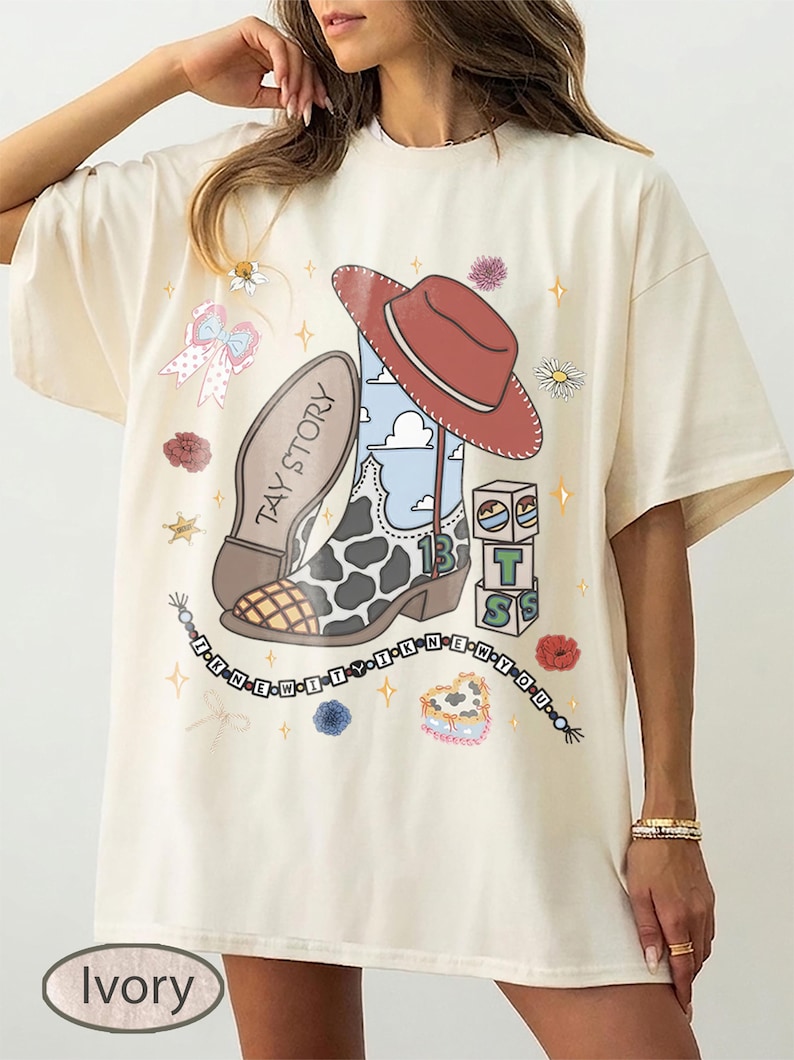 May include: An ivory t-shirt featuring a graphic print of a cowboy boot with a cow print pattern, a red cowboy hat, alphabet blocks, and a beaded string with the text I KNEW IT. The design includes small flowers, stars, and a bow.