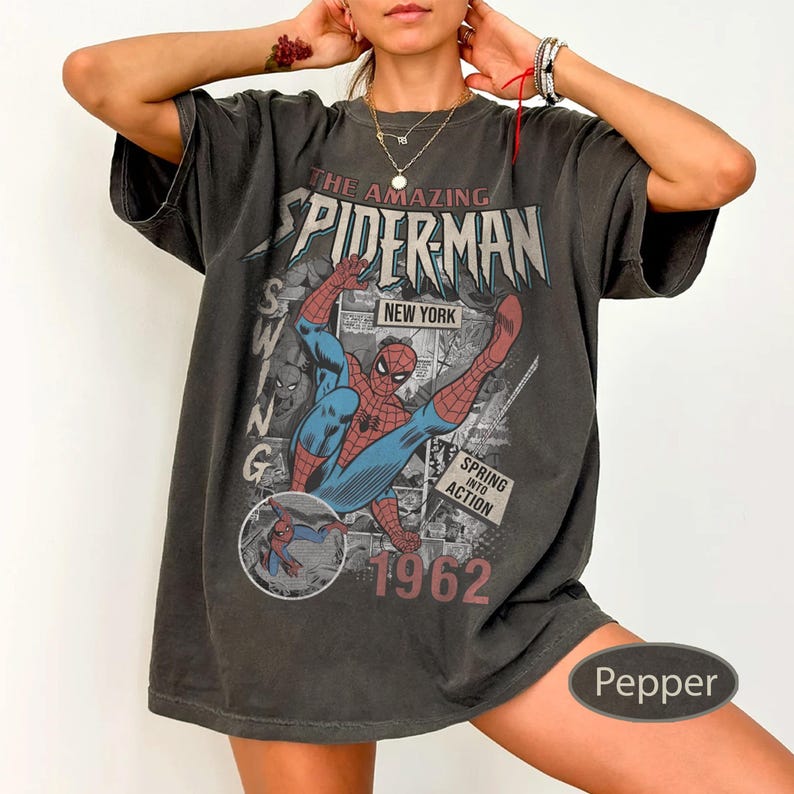 May include: Dark gray t-shirt featuring a comic book graphic of Spider-Man in action. The shirt has the text "THE AMAZING SPIDER-MAN NEW YORK 1962" and "SPRING INTO ACTION". The shirt is oversized and has short sleeves.