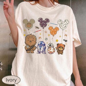 May include: Ivory-colored t-shirt featuring a colorful graphic of Star Wars characters and Mickey Mouse-shaped balloons. The design includes Chewbacca, R2-D2, BB-8, and an Ewok, with balloons in various colors and patterns. The shirt has a relaxed fit.