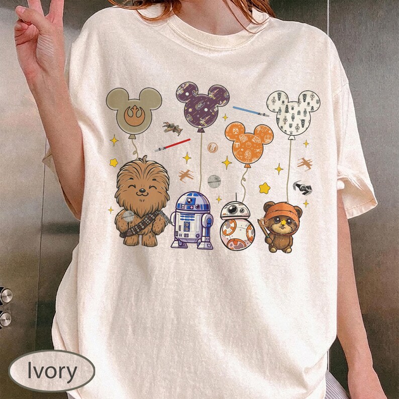 May include: Ivory-colored t-shirt featuring a colorful graphic of Star Wars characters and Mickey Mouse-shaped balloons. The design includes Chewbacca, R2-D2, BB-8, and an Ewok, with balloons in various colors and patterns. The shirt has a relaxed fit.