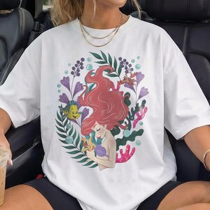 Peut inclure: T-shirt blanc avec une illustration color&eacute;e d'une sir&egrave;ne aux cheveux roux, entour&eacute;e de plantes aquatiques et d'un poisson jaune. Le motif comprend des accents violets, verts et corail, cr&eacute;ant une esth&eacute;tique fantaisiste et artistique. Le t-shirt est de style d&eacute;contract&eacute; et surdimensionn&eacute;.