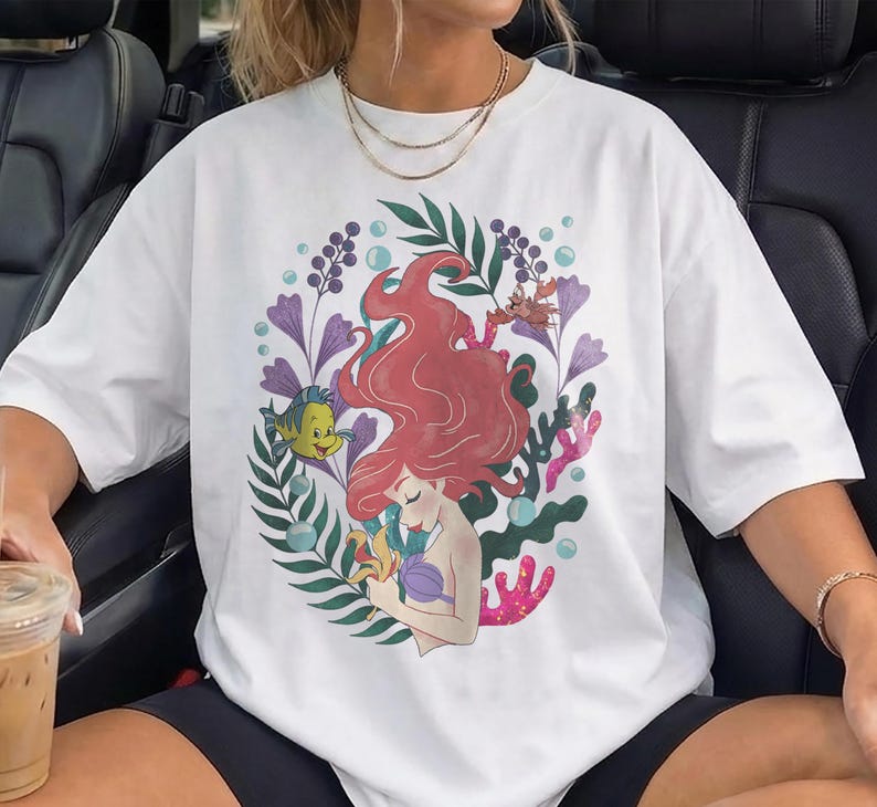 Peut inclure: T-shirt blanc avec une illustration color&eacute;e d'une sir&egrave;ne aux cheveux roux, entour&eacute;e de plantes aquatiques et d'un poisson jaune. Le motif comprend des accents violets, verts et corail, cr&eacute;ant une esth&eacute;tique fantaisiste et artistique. Le t-shirt est de style d&eacute;contract&eacute; et surdimensionn&eacute;.